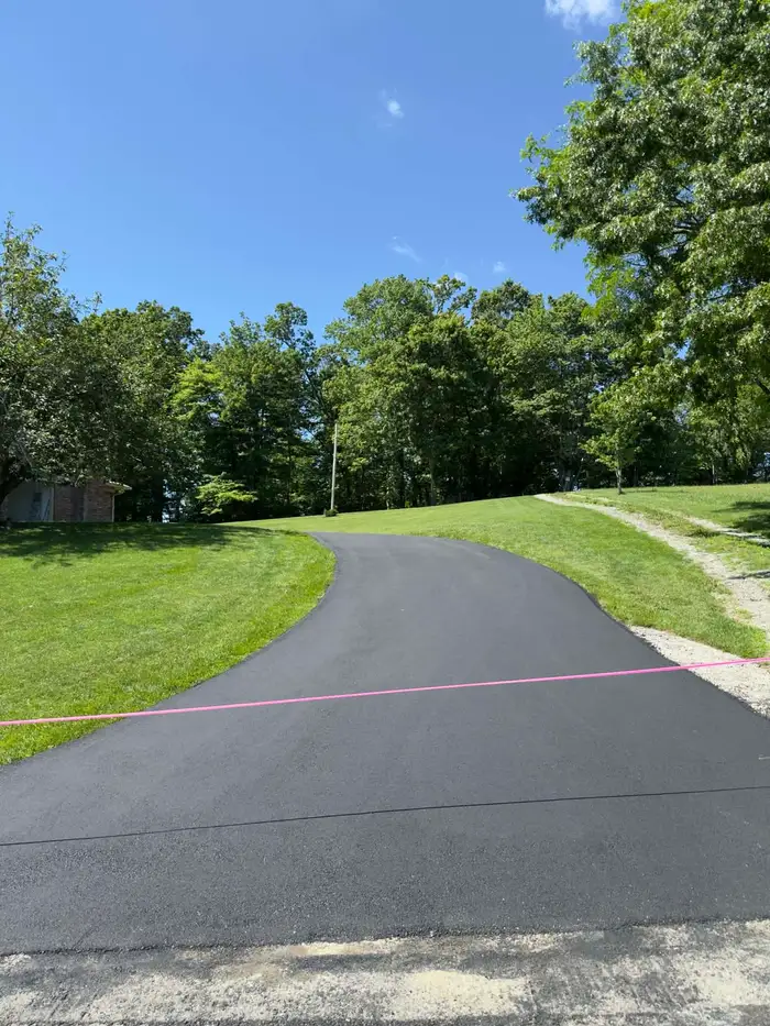 New asphalt driveway curving uphill across a mown lawn, taped off while the surface cures