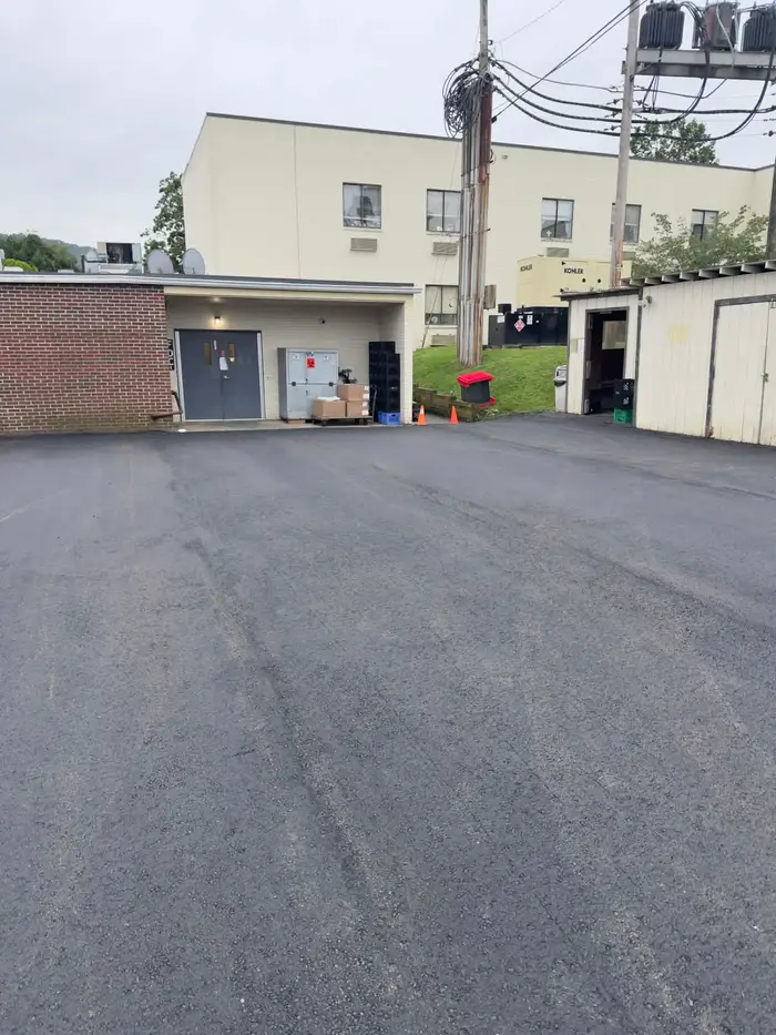 Fresh asphalt across a delivery yard behind a large building, with loading doors and stacked crates
