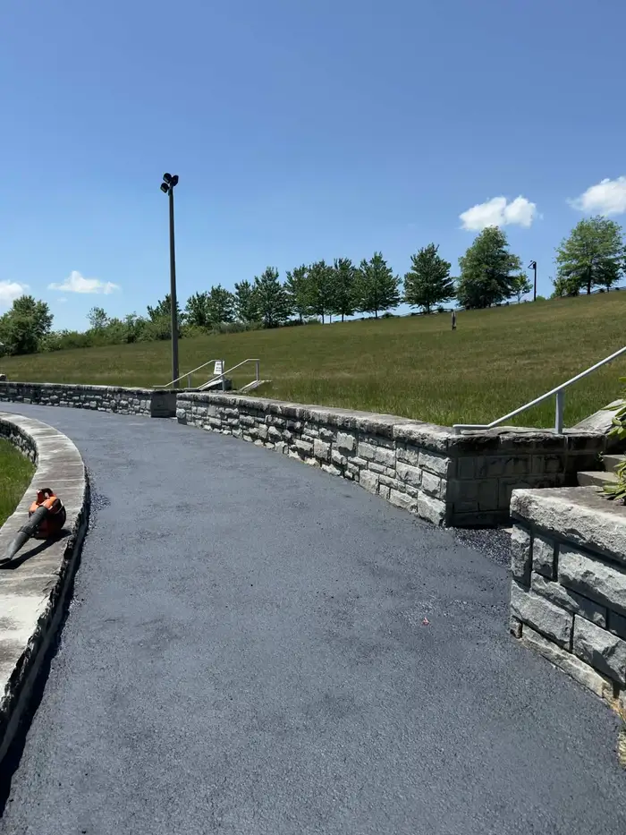 Curved asphalt drive running beside a stacked stone retaining wall, leaf blower resting on the kerb