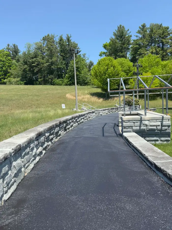 Freshly sealed drive curving past a stone retaining wall towards a steel canopy frame on a terrace