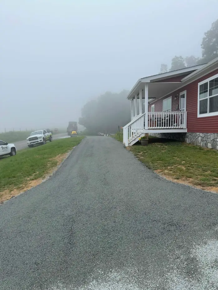 Newly laid tar and chip driveway beside a red sided house on a foggy morning, work trucks on the road