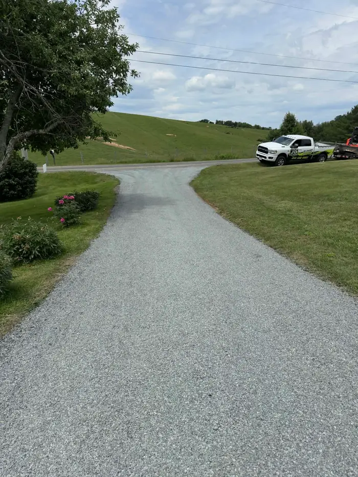 Tar and chip driveway flaring out to meet a rural road, pasture hill behind and the Lines by JD truck parked up
