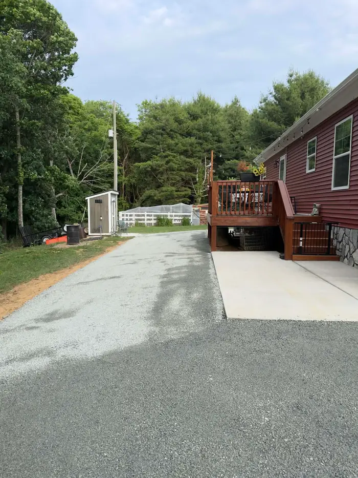 Tar and chip drive and parking area running past a red house with a wooden deck and concrete patio
